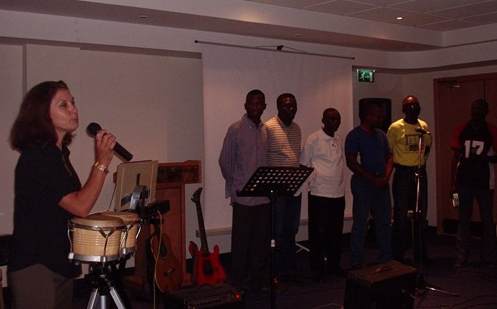 6 men lined up in the front of a hall, and a lady holding a microphone standing next to a set of bongo drums.